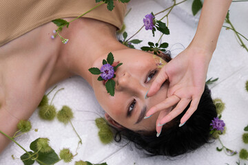 Young woman lying supine on a surface covered with plants, holding a purple flower near her face."