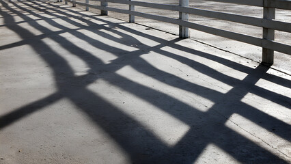 A series of fence rails forming strong parallel shadows across a slightly textured concrete floor in harsh sunlight