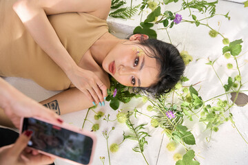 Overhead view of a young woman holding a smartphone, lying on a bed of green plants and purple flowers."
