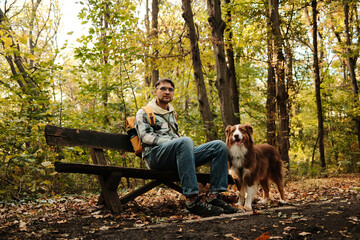 Man with Australian Shepherd dog sitting on old bench in sunlit forest, enjoying nature. Concept of freedom, rest, and companionship