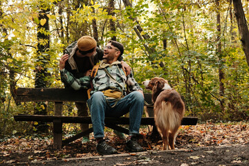 Two friends or couple with Australian Shepherd dog sharing laughter on park bench surrounded by autumn trees. Concept of friendship and joy