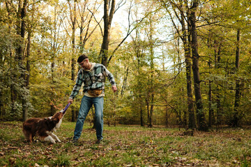 Man and Australian Shepherd dog playing tug-of-war with puller toy in autumn forest. Concept of activity, training, and friendship.