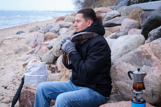 Middle age brunette man making coffee on a cold seaside beach during family outdoors. Winter lifestyle scenes with warm clothing, coastal environment and everyday activities.