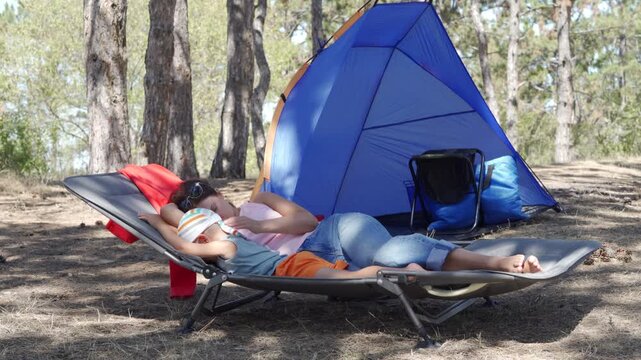 A mother and son lie together on a camping cot. The boy rests on his mom while she relaxes near a tent. They enjoy a quiet moment of bonding in the sunny pine forest.