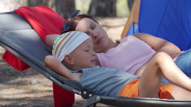 A mother and son lie together on a camping cot. The boy rests on his mom while she relaxes near a tent. They enjoy a quiet moment of bonding in the sunny pine forest.