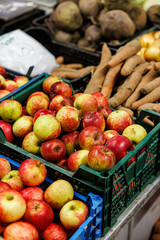 Red and green apples stacked in plastic crates on a produce stall, surrounded by carrots and beets in an indoor market setting