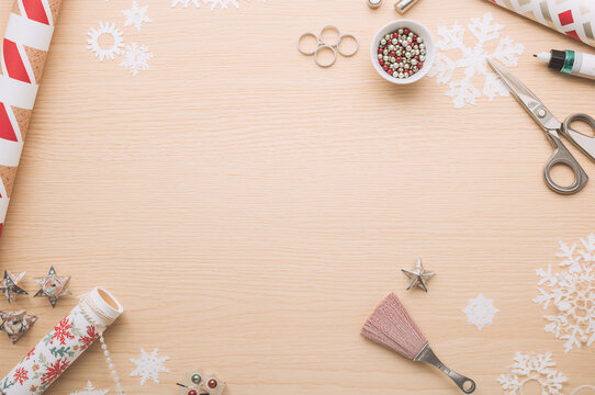 Light wooden background framed by wrapping paper rolls, scissors, snowflake cutouts, and decorations, symbolizing Christmas gift wrapping and holiday crafting preparation. copy space - Powered by Adobe