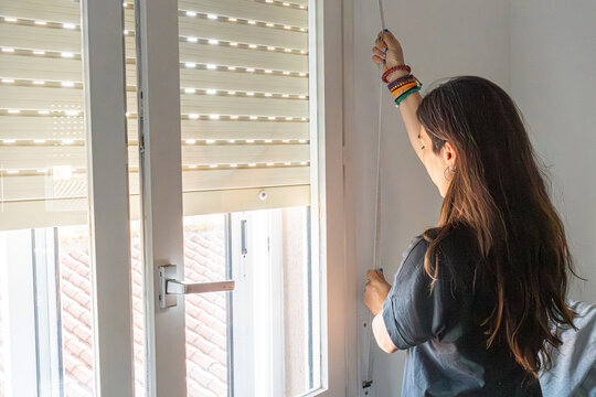 Woman adjusting roller blind on window, controlling light and privacy in a domestic setting