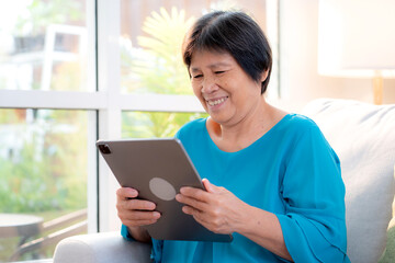 Senior asian woman relax on sofa reading tablet in living room at home, online connection and digital lifestyle, elder watching tablet for social media, remote learning, elderly using chatbot or AI.