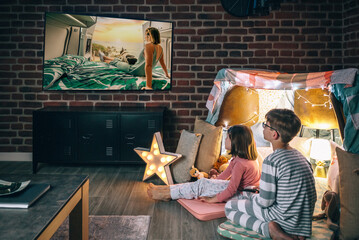 Two concentrated children sitting over mats while watching television program on living room