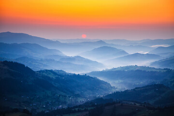 Breathtaking view from above captures the sunset behind layers of mountains surrounded by soft mist in the valleys, creating a calm atmosphere at dusk. Photo wallpaper. Discovery the beauty of earth.