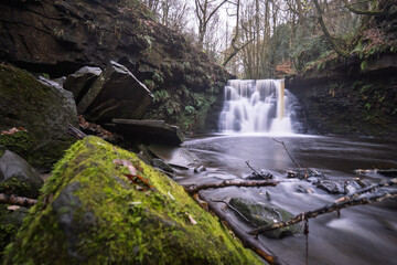 Long-exposure of a tranquil woodland waterfall on an autumn day. Moss and leaf covered rocks, soft flowing water, and rich forest textures create a peaceful natural landscape ideal for outdoor themes.
