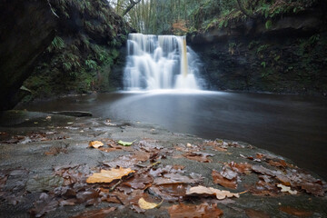 Long-exposure of a tranquil woodland waterfall on an autumn day. Moss and leaf covered rocks, soft flowing water, and rich forest textures create a peaceful natural landscape ideal for outdoor themes.