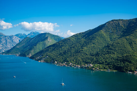 Aerial view of the Bay of Kotor in Montenegro. Lush green mountains rise above the calm blue waters, dotted with small boats and a clear blue sky.
