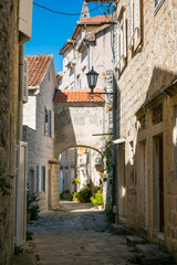 Narrow cobblestone street in Perast, Montenegro. Stone buildings with red roofs line the path. A vintage street lamp hangs above, creating a charming atmosphere.