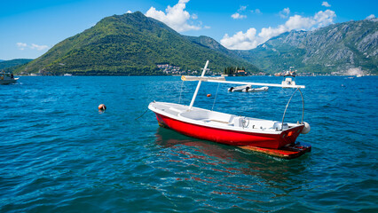 A red boat floats on clear blue water in Montenegro, surrounded by green mountains under a bright sky. The scene captures the beauty of the Balkans.