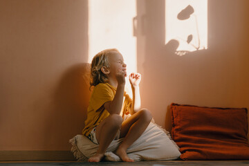 Cutest little boy smiling and squinting in sunlight in front of wall indoors. Happy child looking at sun in window at home. Portrait of playful child preschool age. Childhood lifestyle photography.