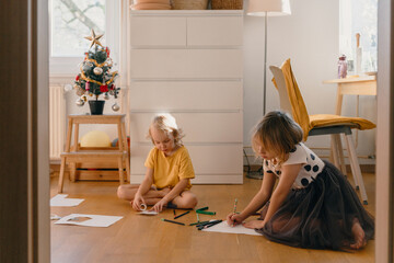 Two children sit on floor drawing on paper in living room decorated with small Christmas tree. Kids being creative while playing at home. Younger brother with older sister together indoors.