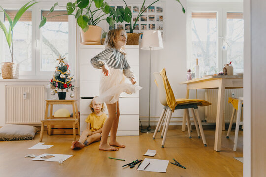 Playful girl twirls in her dress while her younger brother sits on the floor in a messy living room decorated with a small Christmas tree. Warm and cheerful family moments at home.