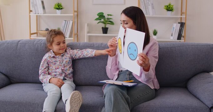 Female young psychologist showing cards with positive and negative emotions faces to little cute girl during therapy session in office. Development of emotional intelligence concept. 4k video.