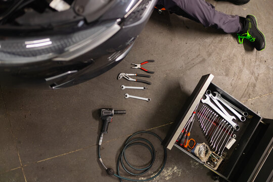 Mechanic under car with tools on floor: wrenches, pliers, impact gun and tool chest. Auto repair in professional workshop.