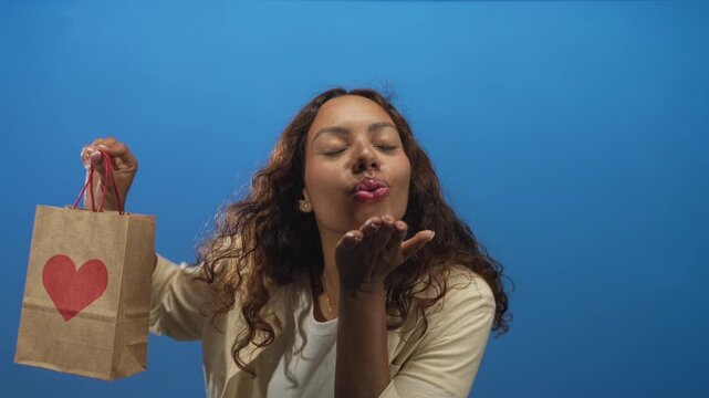 Young african american woman holding heart printed gift bag and blowing a kiss toward camera in a bright blue studio setting; playful love.