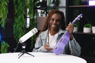 Happy Joyful african female adult holding purple ukulele in studio with microphone on online video lesson streaming broadcast