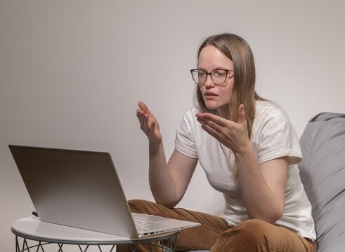 A young woman wearing glasses speaks during an online call, gesturing while working on her laptop in