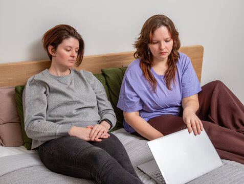 Two young women relax on a bed together, opening a laptop to start a movie or series while enjoying