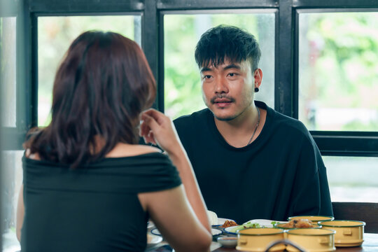 Asian adult man discussing business plan with woman in small restaurant during lunch, food served on wooden table, cafe interior with daylight window, teamwork, communication, partnership