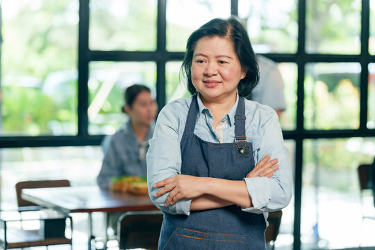 Asian elderly woman small business owner in apron smiling proudly while standing inside cafe representing confidence experience teamwork dedication in restaurant management and hospitality lifestyle - Powered by Adobe