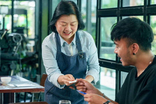 Asian female restaurant owner smiles while accepting credit card from male customer for contactless payment inside bright cafe small business scene representing modern cashless lifestyle service