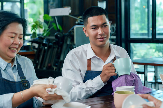 Asian young male barista holding mug chatting with senior female owner while cleaning dishes teamwork concept cafe restaurant small business cooperation family hospitality management friendly