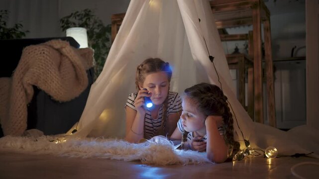 Girls explore book with flashlight inside cozy tent lying on rug under stringlight and blanket while reading story during nighttime play showing sister friendship imagination pajama soft warm glow