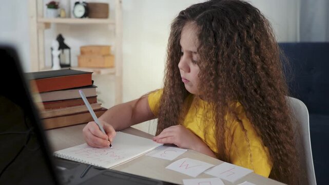 Girl studying with laptop at desk focused on homework and writing notes while arranging flashcards for school learning and concentration child practicing spelling and study routine with attention
