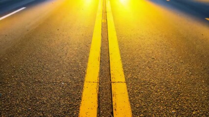 Close-up view of a smooth asphalt road with yellow lines captured during a transition from daytime to evening highlighting bright yellow headlights illuminating the surface.