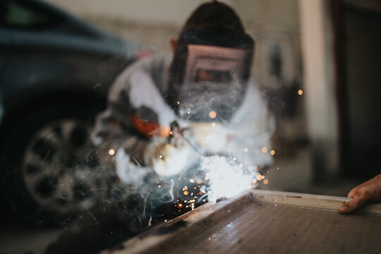 A close-up of a welder in safety gear shaping metal at a workbench in a garage, with bright sparks and smoke capturing skilled fabrication and industrial craft.