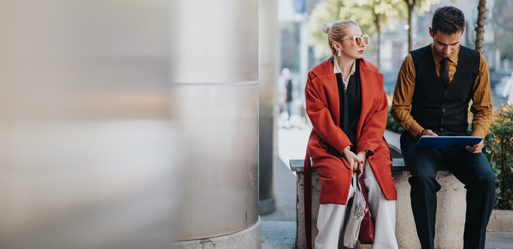Two well-dressed professionals sit on a city bench outdoors. A woman in a red coat and a man in a vest discuss work as they glance at a tablet, conveying a moment of urban collaboration.
