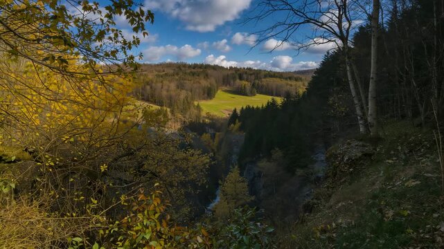 Scenic view of a lush forest valley with autumn foliage gentle flowing river and rolling hills under a vibrant blue sky with scattered clouds in natural daylight.