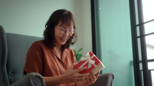 Happy pretty senior asian woman exciting while opening surprised a red gift box with joyful holiday celebration, healthy aging retirement lifestyle gratitude love and festive happiness concept.
