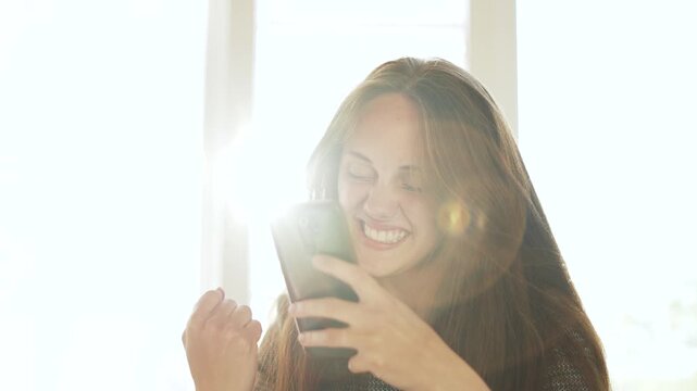 Woman using smartphone and smiling while texting by sunlight window with lens flare, long hair framing face, hand holding device, closeup portrait showing relaxed expression and natural light on skin