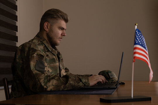  A soldier is working on his laptop at a desk with an American flag nearby.