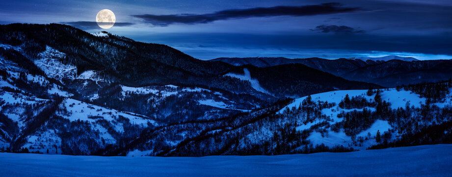 panorama with snowy forested hills in winter at night. beautiful landscape of mountain ridge with snow covered peaks in the distance in full moon light. backdrop for fiction art or mystery concepts