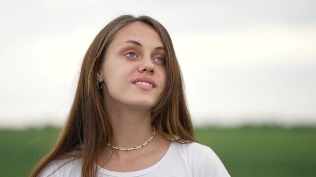 Young woman stands in field with long hair and shell necklace, eyes closed then smiling, wearing white shirt, green grass and soft cloudy sky behind, calm portrait of contemplative young adult