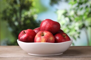 Ripe red apples in bowl on wooden table against blurred green background, closeup