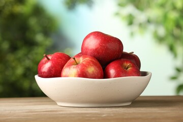 Ripe red apples in bowl on wooden table against blurred green background, closeup