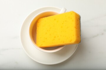 Clean kitchen sponge in cup and saucer on white marble table, top view