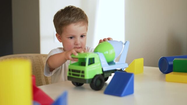 Boy plays with toy truck on table indoor in preschool room, with concentration he manipulates plastic cement mixer and block, engaged in focused play and toy exploration with careful hand movement