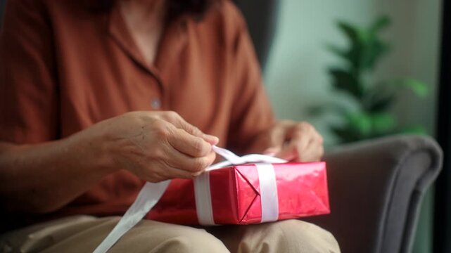 Cropped shot hands of senior woman tying white ribbon on a red gift box at home for holiday celebration, healthy retirement lifestyle gratitude love and festive happiness concept.
