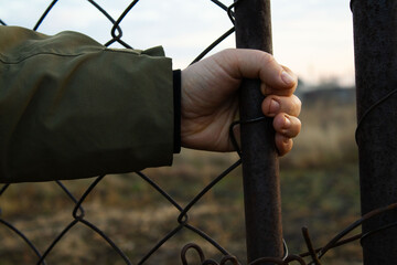Hand in a Khaki Sleeve Grasping a Rusted Metal Fence Post with Wire Mesh